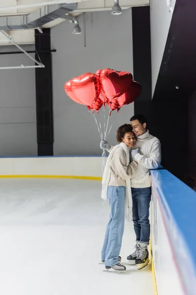 Веселая межрасовая пара, держащаяся за руки и воздушные шары в форме сердца на катке — стоковое фото