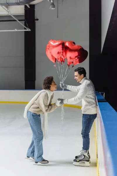 Vista lateral de una mujer afroamericana positiva cogida de la mano de un novio con globos en forma de corazón en una pista de hielo - foto de stock