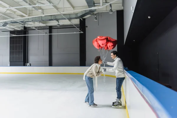 Side view of smiling man holding heart shaped balloons and hand of african american girlfriend on ice rink — Stock Photo
