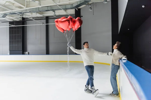 Cheerful african american woman holding hand of boyfriend with heart shaped balloons on ice rink — Stock Photo