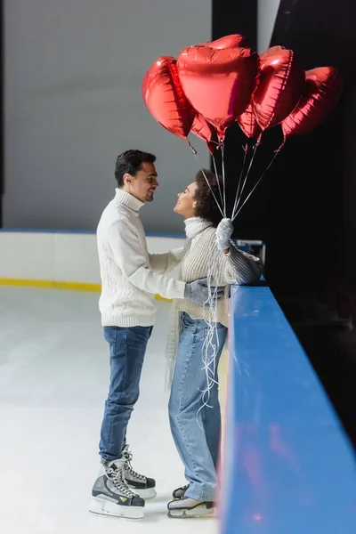 Vista lateral del joven en patines de hielo abrazando a la novia afroamericana sosteniendo globos en forma de corazón en la pista - foto de stock