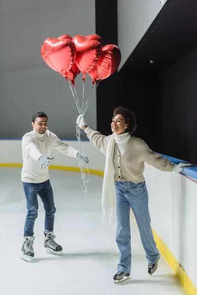Femme afro-américaine souriante tenant des ballons rouges en forme de coeur tandis que le patinage sur glace près de petit ami sur la patinoire — Photo de stock
