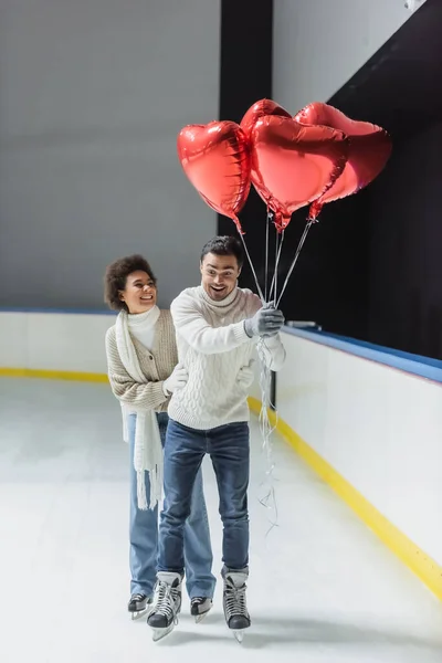 Positive multiethnic couple with heart shaped balloons ice skating on rink — Stock Photo