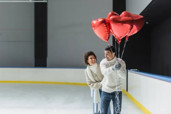 Sorrindo afro-americano mulher abraçando namorado com balões em forma de coração na pista de gelo — Fotografia de Stock