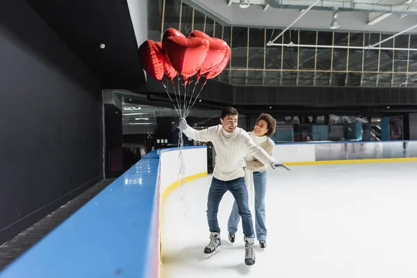Mulher americana africana alegre em roupas quentes patinação no gelo perto do namorado com balões em forma de coração na pista — Fotografia de Stock