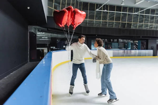 Positive interracial couple in warm sweaters holding heart shaped balloons while ice skating on rink — Stock Photo