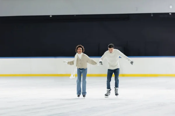Sonriente pareja multiétnica en suéteres tomados de la mano mientras patina sobre hielo en la pista - foto de stock