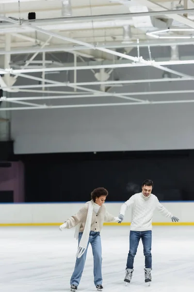 Sonriente pareja multiétnica en guantes y suéteres patinaje sobre hielo en pista - foto de stock