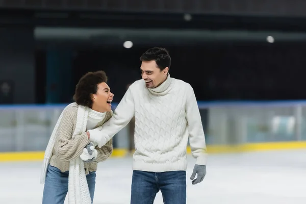 Cheerful african american woman holding hand of boyfriend in sweater and gloves on ice rink — Stock Photo