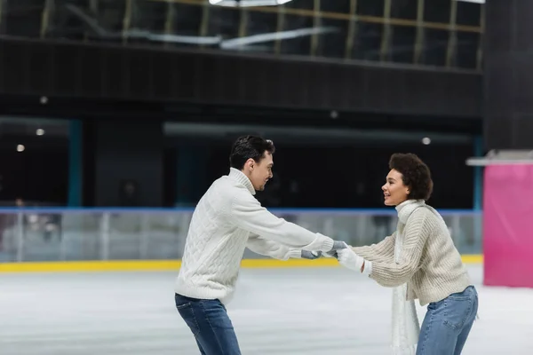Vista lateral de una joven pareja interracial cogida de la mano mientras patina sobre hielo en la pista de patinaje - foto de stock