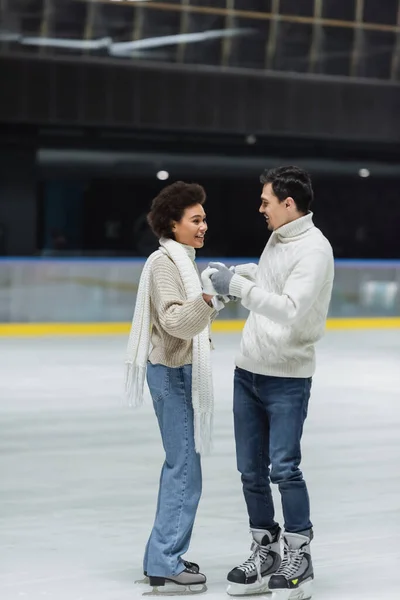 Joven en suéter cogido de la mano de la novia afroamericana en guantes en pista de hielo - foto de stock