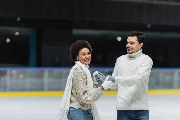 Cheerful african american woman looking at camera and holding hands of boyfriend on ice rink — Stock Photo