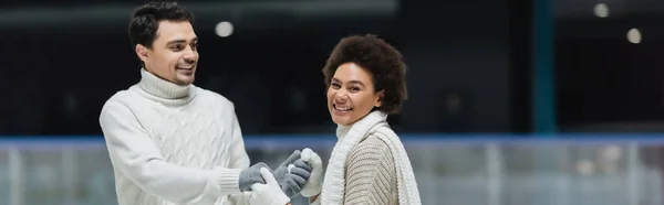 Young african american woman smiling at camera and holding hands of boyfriend on ice rink, banner — Stock Photo