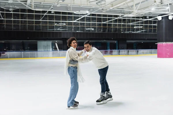 Couple multiethnique positif dans des gants regardant la caméra tout en s'amusant sur la patinoire — Photo de stock