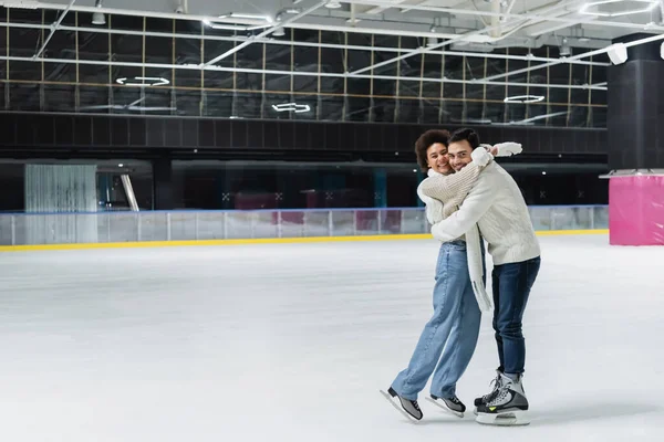 Multiethnic couple in warm clothes embracing and looking at camera on ice rink — Stock Photo