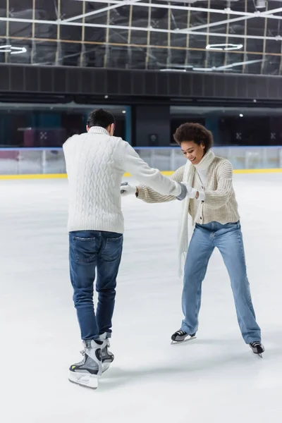 Sonriente mujer afroamericana cogida de la mano del novio en patines de hielo en la pista de patinaje - foto de stock
