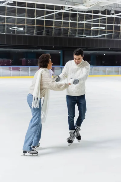 Couple interracial joyeux en jeans et pulls tenant la main pendant le patin à glace sur la patinoire — Photo de stock