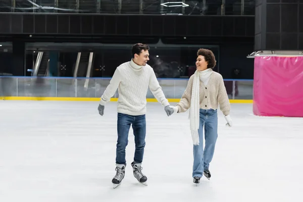 Cheerful interracial couple in gloves and sweaters skating on ice rink — Stock Photo