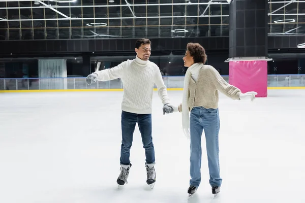 Sonriente pareja interracial en guantes y suéteres tomados de la mano en pista de hielo - foto de stock