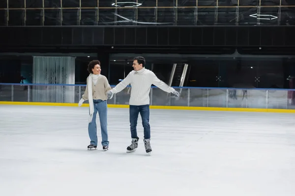 Positivo interracial pareja de patinaje sobre hielo durante la fecha en pista de patinaje - foto de stock