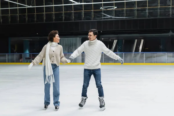Sonriente mujer afroamericana en jeans y suéter patinaje sobre hielo con novio en pista de patinaje - foto de stock