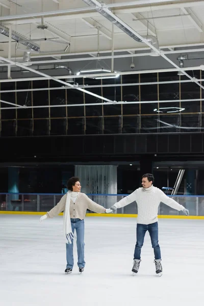 Sorrindo jovem casal interracial patinação no gelo e de mãos dadas na pista — Fotografia de Stock