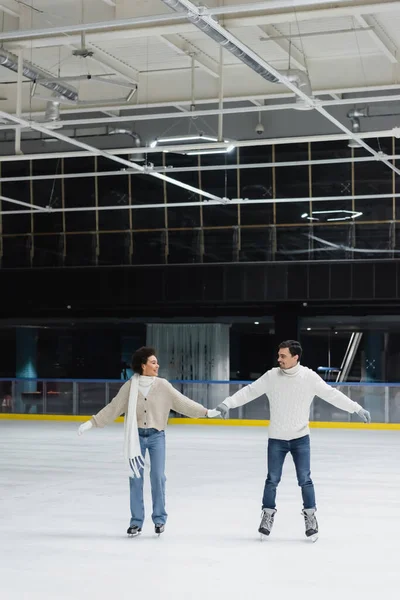 Sonriente joven interracial pareja de patinaje sobre hielo y de la mano en la pista de patinaje — Stock Photo