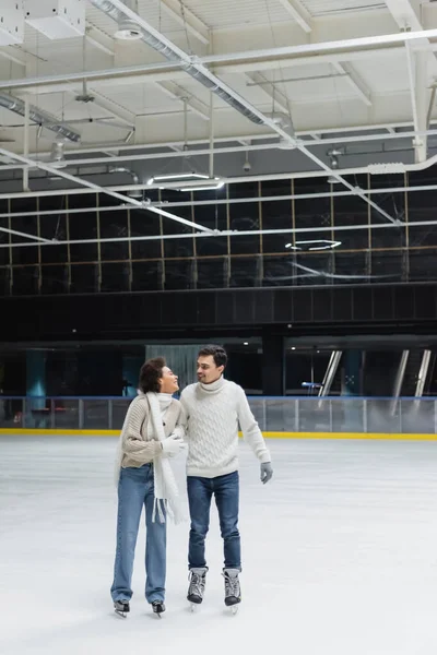 Jeune femme afro-américaine en pull et jeans patinage sur glace et en regardant petit ami sur la patinoire — Photo de stock