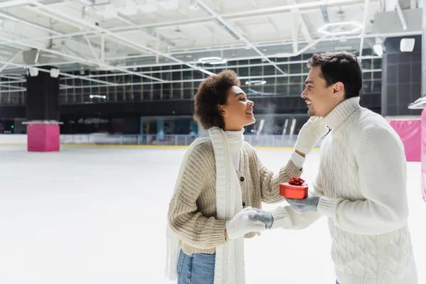 Side view of smiling man holding heart shaped gift box and hand of african american girlfriend on ice rink — Stock Photo