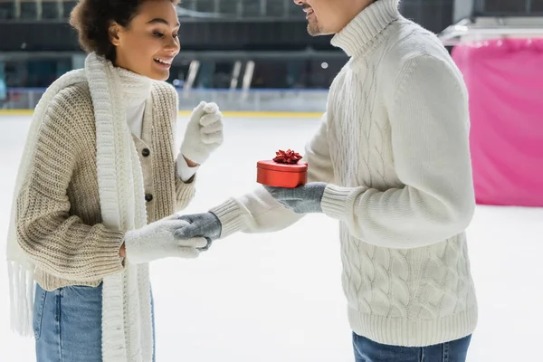 Homem sorridente segurando caixa em forma de coração e mão de namorada afro-americana na pista de gelo — Fotografia de Stock