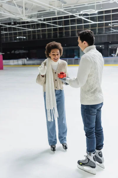 Sorrindo Africano americano mulher olhando para coração em forma de presente perto namorado na pista de gelo — Fotografia de Stock