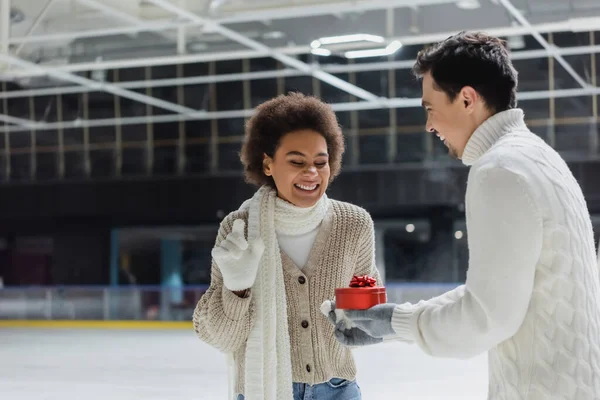 Cheerful man holding heart shaped gift near excited african american girlfriend on ice rink — Stock Photo