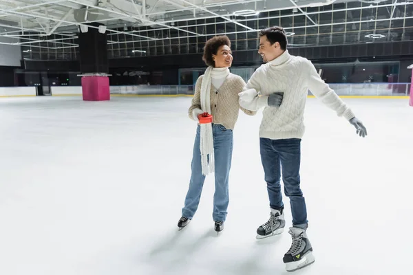 Smiling african american woman holding gift box and looking at boyfriend on ice rink — Stock Photo