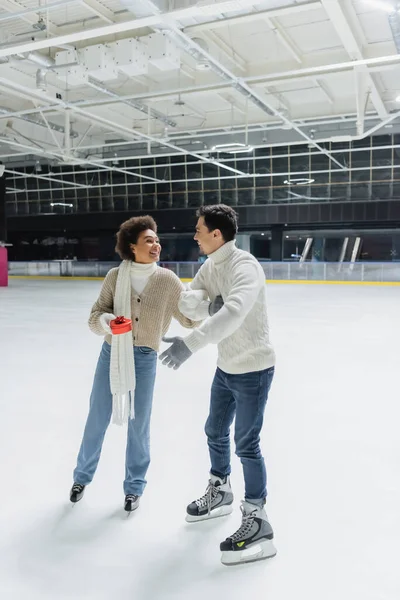 Positive african american woman holding red gift and looking at boyfriend on ice rink — Stock Photo