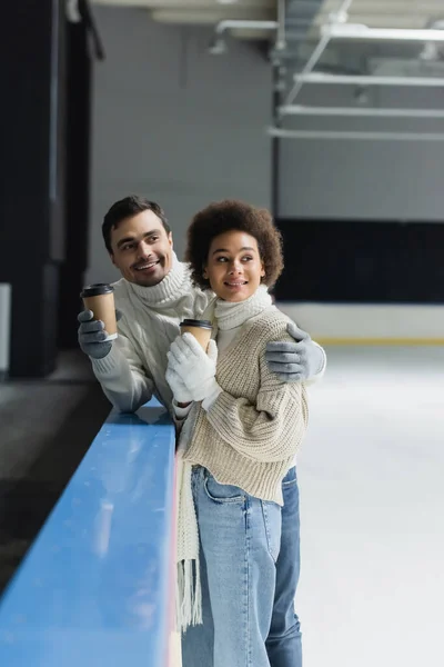 Hombre sonriente abrazando novia afroamericana con café para ir en pista de hielo - foto de stock