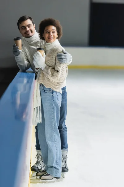 Uomo sorridente che abbraccia la ragazza africana americana con tazza di carta sulla pista di pattinaggio sul ghiaccio — Foto stock