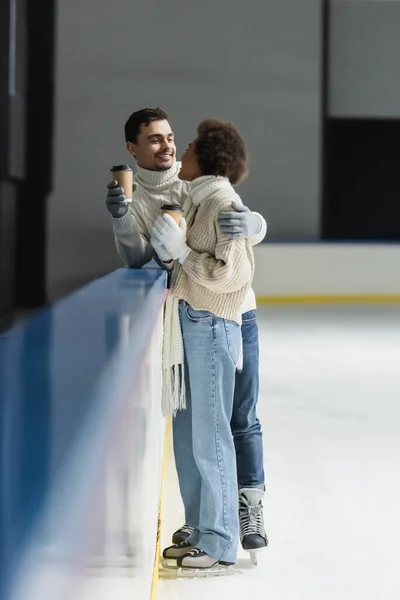 Jovem de luvas segurando café para ir e abraçando a namorada afro-americana no ringue de gelo — Fotografia de Stock