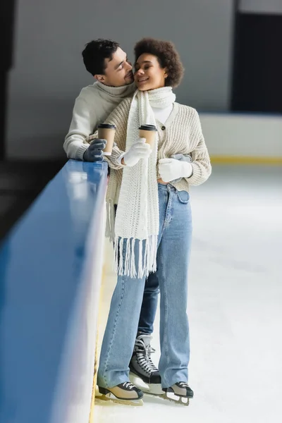 Smiling man kissing and hugging african american girlfriend with coffee to go on ice rink — Stock Photo
