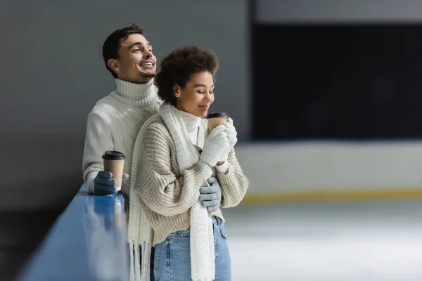 Couple interracial positif dans des gants tenant du café pour aller sur la patinoire — Photo de stock