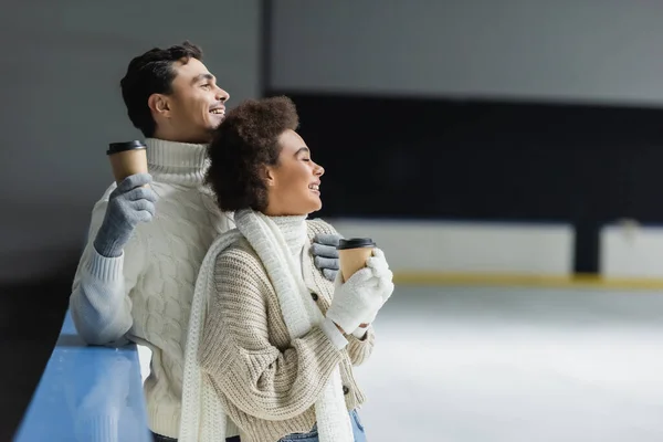 Vista lateral de alegre pareja interracial sosteniendo café para ir y mirando hacia otro lado en pista de hielo - foto de stock