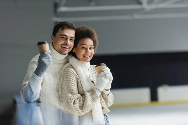 Alegre casal interracial em roupas quentes segurando café para ir e olhando para a câmera na pista de gelo — Fotografia de Stock