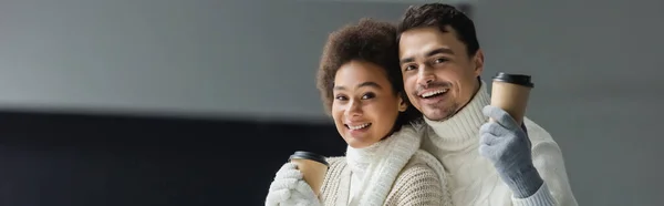 Positives multiethnisches Paar in warmer Kleidung mit Pappbechern und Blick in die Kamera, Banner — Stockfoto