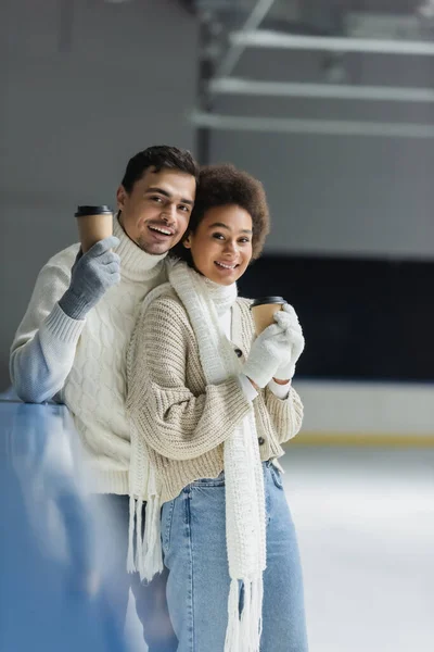 Alegre interracial casal segurando takeaway bebida e olhando para câmera no gelo rink — Fotografia de Stock