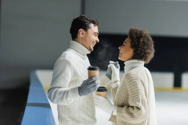 Vista lateral de la sonriente pareja interracial cogida de la mano y café para ir en pista de hielo - foto de stock