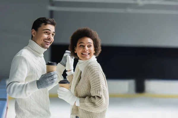 Positive multiethnic couple holding hands and coffee to go on ice rink — Stock Photo