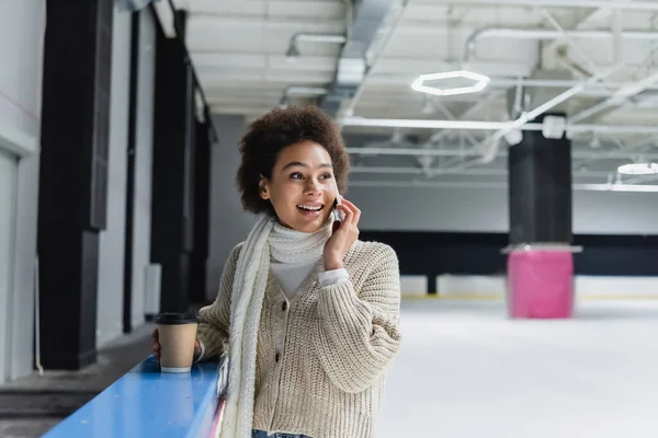 Femme afro-américaine positive parlant sur smartphone et tenant une tasse en papier sur une patinoire — Photo de stock