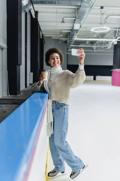 Donna afroamericana in pattinaggio su ghiaccio che tiene il caffè per andare a scattare selfie su smartphone sulla pista di pattinaggio — Foto stock