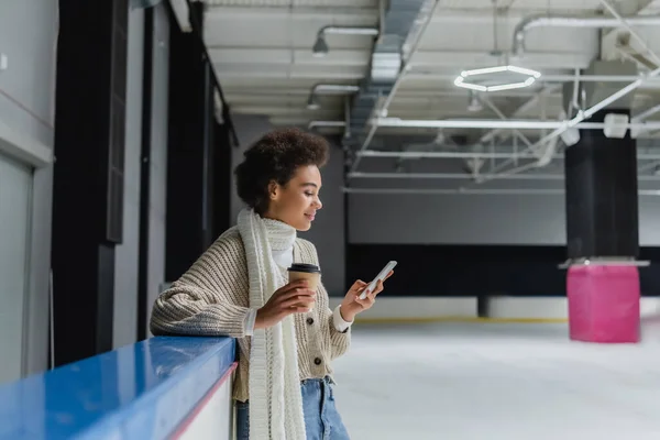Vista lateral de mujer afroamericana en bufanda caliente sosteniendo café para ir y el uso de teléfono celular en pista de hielo - foto de stock