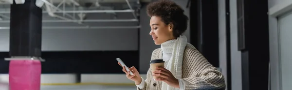 Side view of african american woman holding takeaway coffee and using smartphone on ice rink, banner — Stock Photo