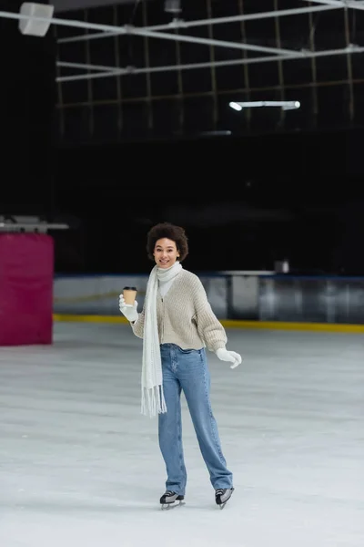 Sonriente mujer afroamericana sosteniendo café para llevar mientras patina sobre hielo en la pista - foto de stock